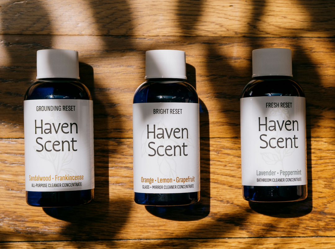 Three small Haven Scent bottles arranged side by side on a wooden surface with strong diagonal shadows. The bottles are dark blue glass with white caps and white labels reading Haven Scent, with scent names Sandalwood-Frankincense, Orange-Lemon-Grapefruit, and Lavender-Peppermint.