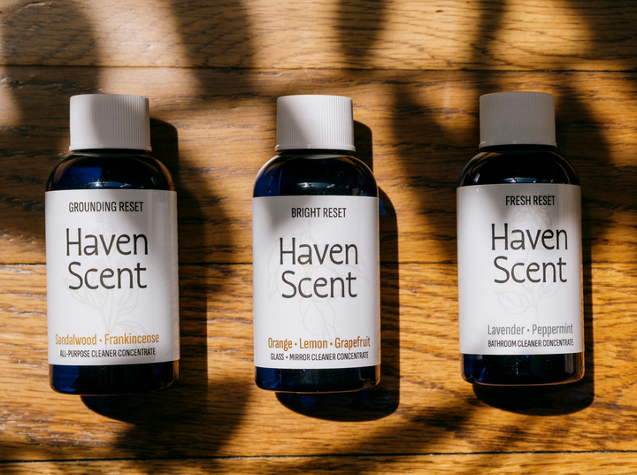 Three small Haven Scent bottles arranged side by side on a wooden surface with strong diagonal shadows. The bottles are dark blue glass with white caps and white labels reading Haven Scent, with scent names Sandalwood-Frankincense, Orange-Lemon-Grapefruit, and Lavender-Peppermint.