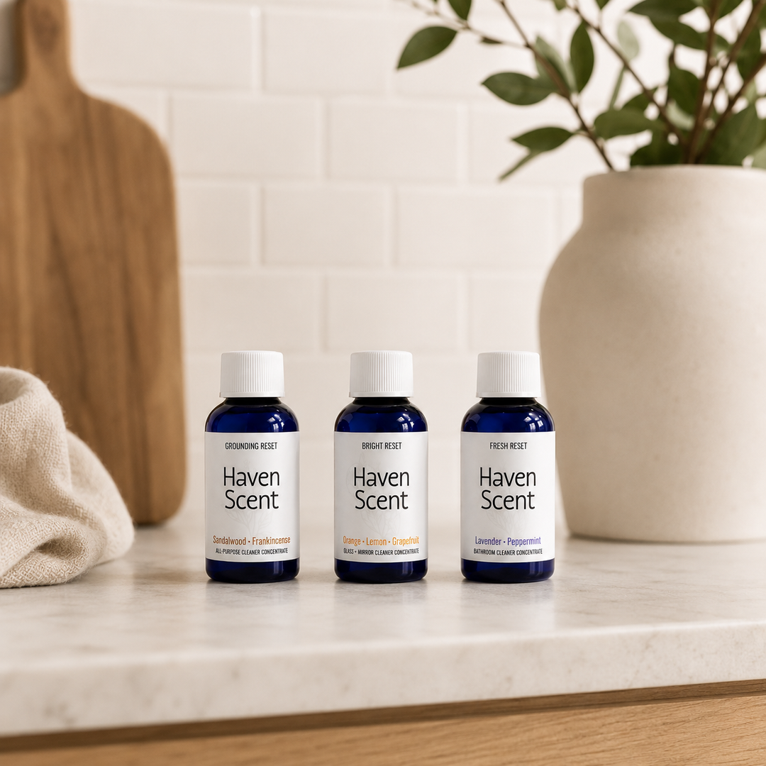 Three small Haven Scent bottles arranged on a light stone countertop in a kitchen-like setting, with a white tile backsplash, a wooden cutting board, a folded beige cloth, and a white vase with greenery in the background.