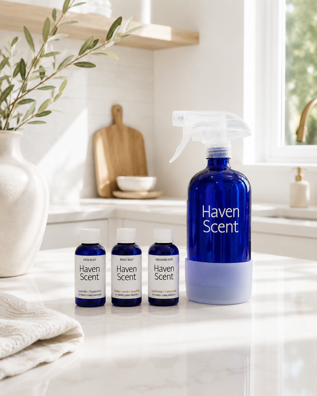 A Haven Scent home fragrance set on a bright white kitchen counter, featuring a large cobalt blue spray bottle with a white base and three matching small spray bottles with white caps.